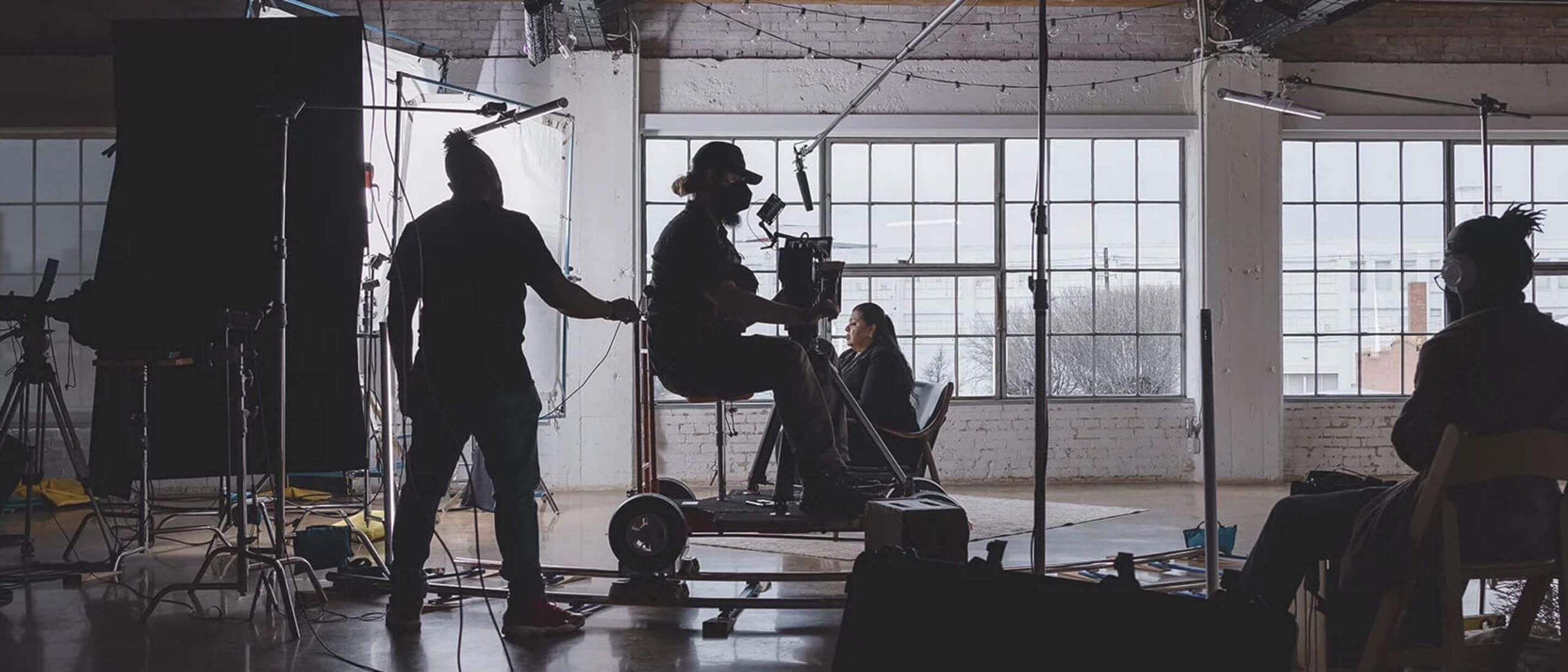 Behind-the-scenes view of a film set with crew members operating a camera on a dolly, capturing an interview scene. The studio features large windows providing natural light, and various lighting and audio equipment is visible, highlighting the production process.